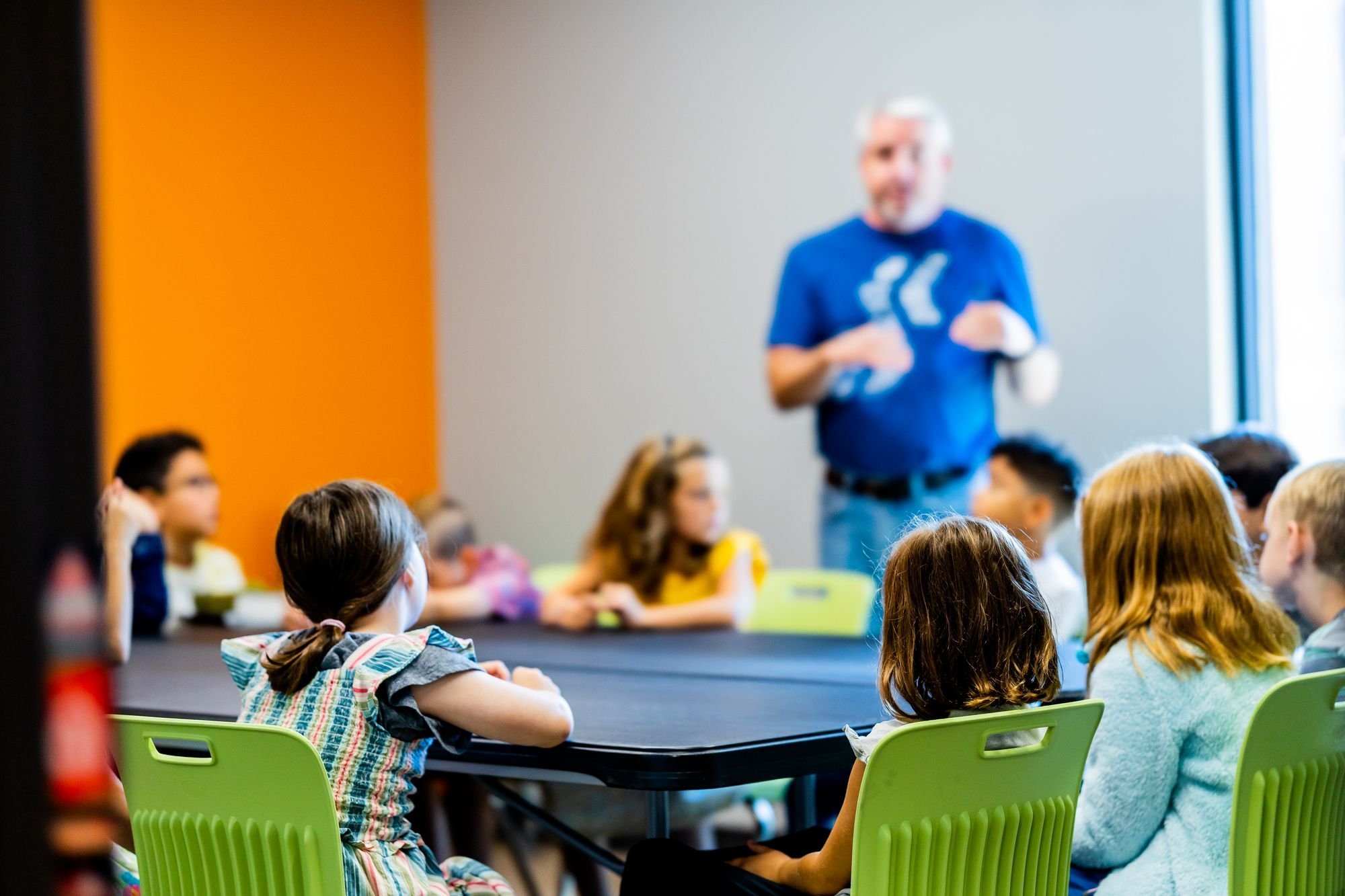 Man in a blue shirt teaching a group of children seated at tables in a classroom