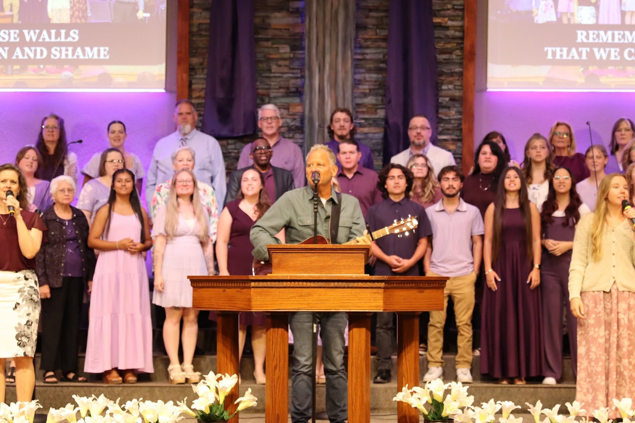 Choir and worship team standing on stage behind a pulpit in a sanctuary with purple lighting